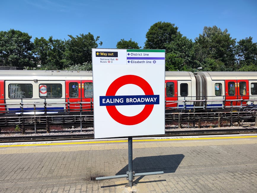 A large white sign with blue and black text displaying 'EALING BROADWAY' is situated on a paved station platform, directly in front of a stationary London Underground train with red, white, and blue exterior. The sign includes directional information, such as a yellow arrow pointing left for the 'Way out,' and mentions the 'National Rail,' 'District line,' and 'Elizabeth line' services. Behind the platform, there are green trees and a clear blue sky. The train is positioned on railway tracks with a metal safety barrier running parallel along the edge of the platform. The scene depicts a typical urban train station environment, which is relevant to home relocation or moving services that involve transportation of furniture and belongings via train or at a transportation hub, often coordinated by companies like Man with Van Ealing during detailed packing and loading processes.