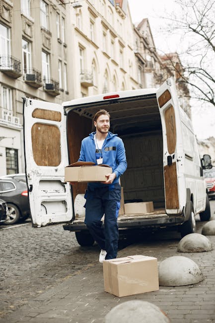 A man wearing a blue uniform standing at the back of an open white van with wooden interior panels, holding a cardboard box in his hands during a home relocation. The van is parked on a cobbled street in an urban residential area with multi-storey buildings and parked cars visible in the background. Additional boxes are placed on the pavement near the van, some on the ground and others being loaded or unloaded. The scene is lit by natural daylight, and the man appears to be engaged in the loading process as part of furniture transport and packing for a move, with the background indicating typical city moving logistics. Man with Van Ealing's services focus on efficient, careful moving solutions in such tight access environments.
