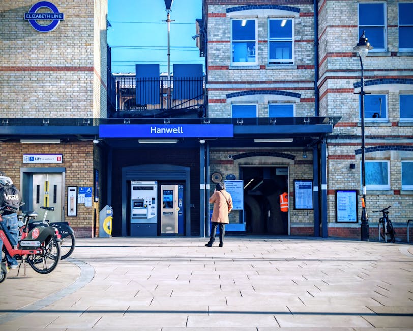 A large white sign with blue and black text displaying 'EALING BROADWAY' is situated on a paved station platform, directly in front of a stationary London Underground train with red, white, and blue exterior. The sign includes directional information, such as a yellow arrow pointing left for the 'Way out,' and mentions the 'National Rail,' 'District line,' and 'Elizabeth line' services. Behind the platform, there are green trees and a clear blue sky. The train is positioned on railway tracks with a metal safety barrier running parallel along the edge of the platform. The scene depicts a typical urban train station environment, which is relevant to home relocation or moving services that involve transportation of furniture and belongings via train or at a transportation hub, often coordinated by companies like Man with Van Ealing during detailed packing and loading processes.
