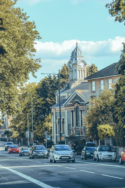 A view of a busy street in Ealing with several parked and moving cars along the pavement. On the left side, mature trees with green foliage line the sidewalk, partially shading the area. In the background, a historic building featuring a prominent clock tower with a pointed spire is visible, surrounded by other structures with traditional brick and stone facades. The scene is illuminated by natural daylight, with a clear blue sky and some scattered clouds overhead. In the context of home relocation or furniture transport, this image depicts a typical urban environment where [COMPANY_NAME], such as Man with Van Ealing, might coordinate the loading process of household goods onto a van for a house removal service, involving careful movement of boxes, furniture, and appliances across the pavement and onto a vehicle, with the backdrop highlighting the regulated area under Ealing Council permit rules for house removals in W5.