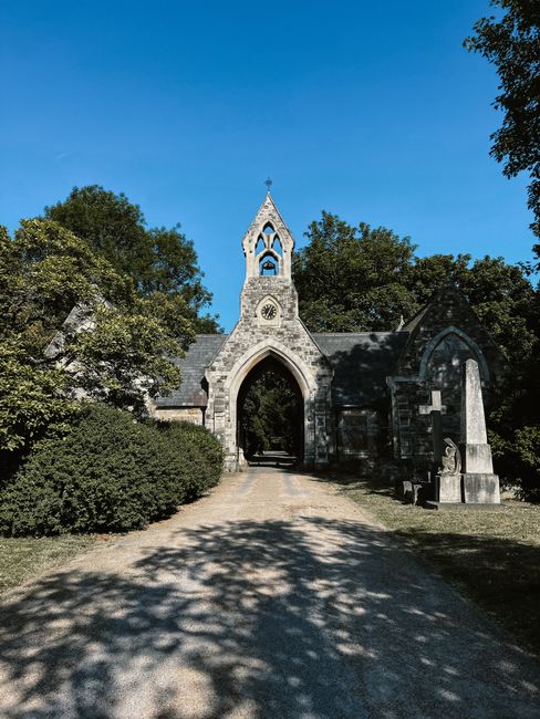 A photograph of a stone church entrance set within a park area during daytime, featuring a tall, pointed archway with a bell tower on top, and a large cross at the peak. The pathway leading to the church gate is lined with neatly trimmed bushes and shadowed by tall trees on both sides. The scene is illuminated by bright sunlight with clear blue skies overhead, creating a contrast between the shaded pathway and the well-lit structure. In the foreground, a gravel walkway is visible, and part of a stone bench is on the right side near the church. This image captures a serene outdoor setting, suitable for house removals and home relocation services as offered by Man with Van Ealing, highlighting the importance of proper access and planning for furniture transport and moving logistics.