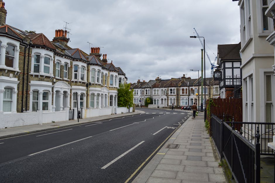 A view of a busy street in Ealing with several parked and moving cars along the pavement. On the left side, mature trees with green foliage line the sidewalk, partially shading the area. In the background, a historic building featuring a prominent clock tower with a pointed spire is visible, surrounded by other structures with traditional brick and stone facades. The scene is illuminated by natural daylight, with a clear blue sky and some scattered clouds overhead. In the context of home relocation or furniture transport, this image depicts a typical urban environment where [COMPANY_NAME], such as Man with Van Ealing, might coordinate the loading process of household goods onto a van for a house removal service, involving careful movement of boxes, furniture, and appliances across the pavement and onto a vehicle, with the backdrop highlighting the regulated area under Ealing Council permit rules for house removals in W5.